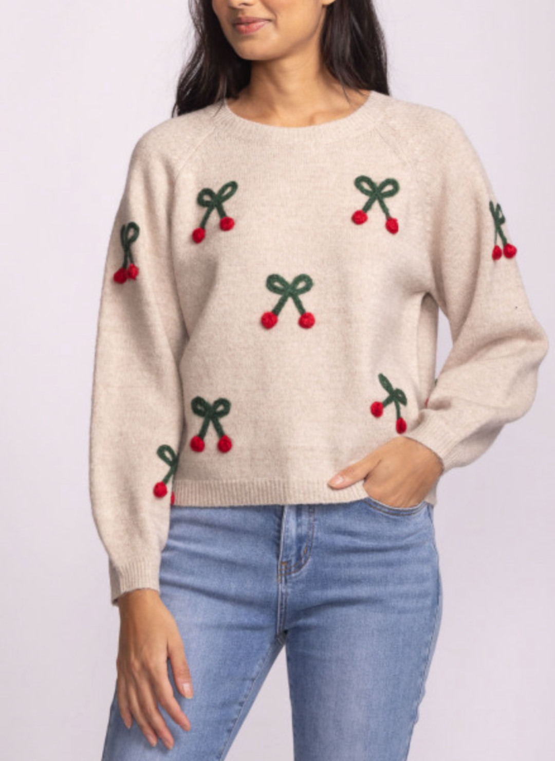Woman wearing the Mon Cherry Sweater from August Boutique Ripon, a soft beige knit with green bow stitching and red pom pom embellishments; features a crew neckline and relaxed fit for cozy, festive holiday style.