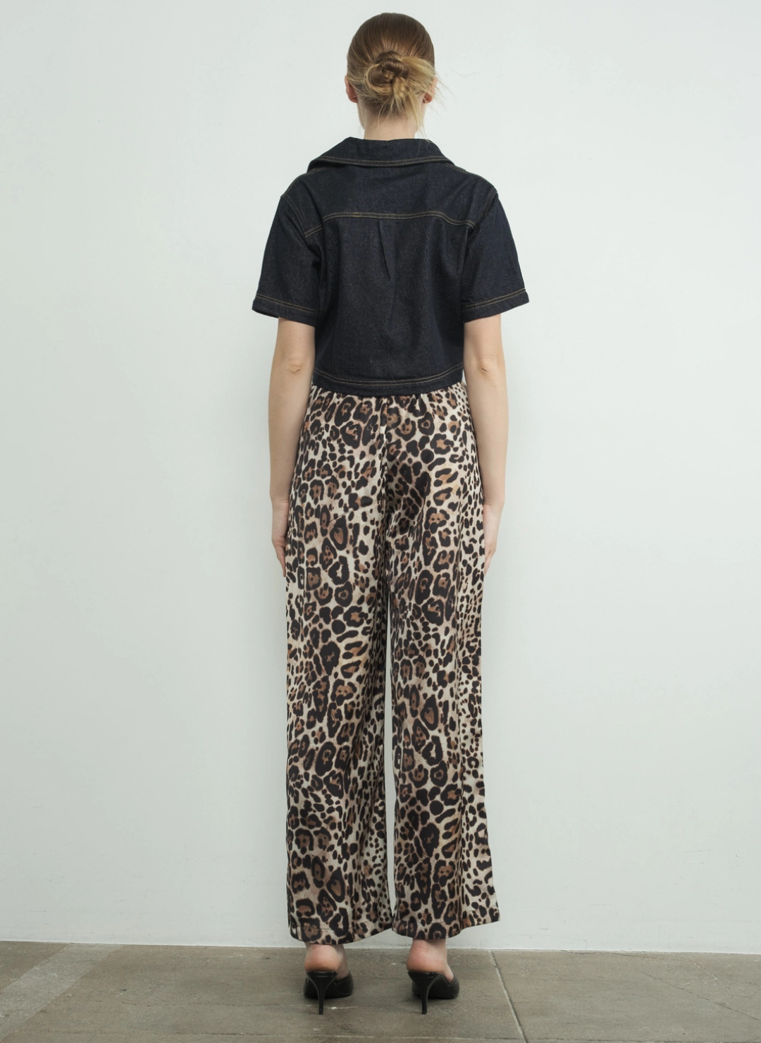 Full back view of model wearing the Dangerously Leopard Satin Pants paired with a black top and black heels. Model is standing infringe of a white background.