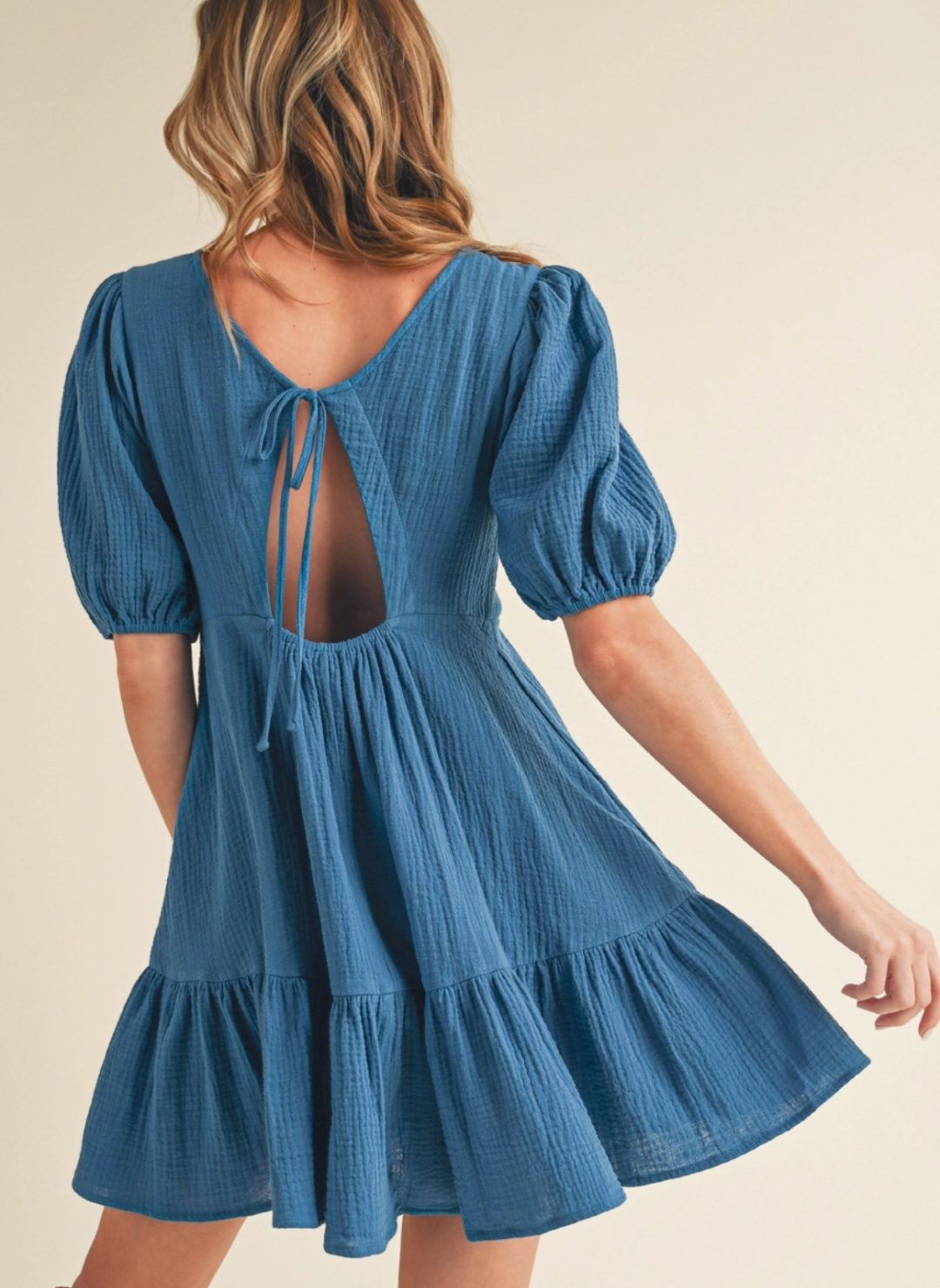 Woman wearing a textured blue mini dress with puff sleeves, open back, and tie detail—featuring a ruffled hem and lightweight gauze fabric, perfect for spring and summer style.