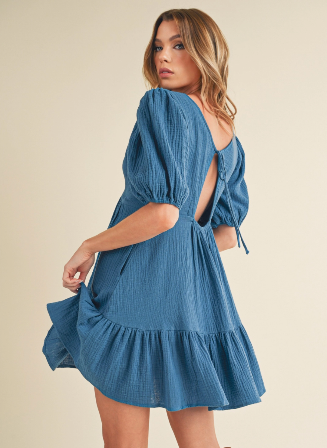 Woman wearing a textured blue mini dress with puff sleeves, open back, and tie detail—featuring a ruffled hem and lightweight gauze fabric, perfect for spring and summer style.