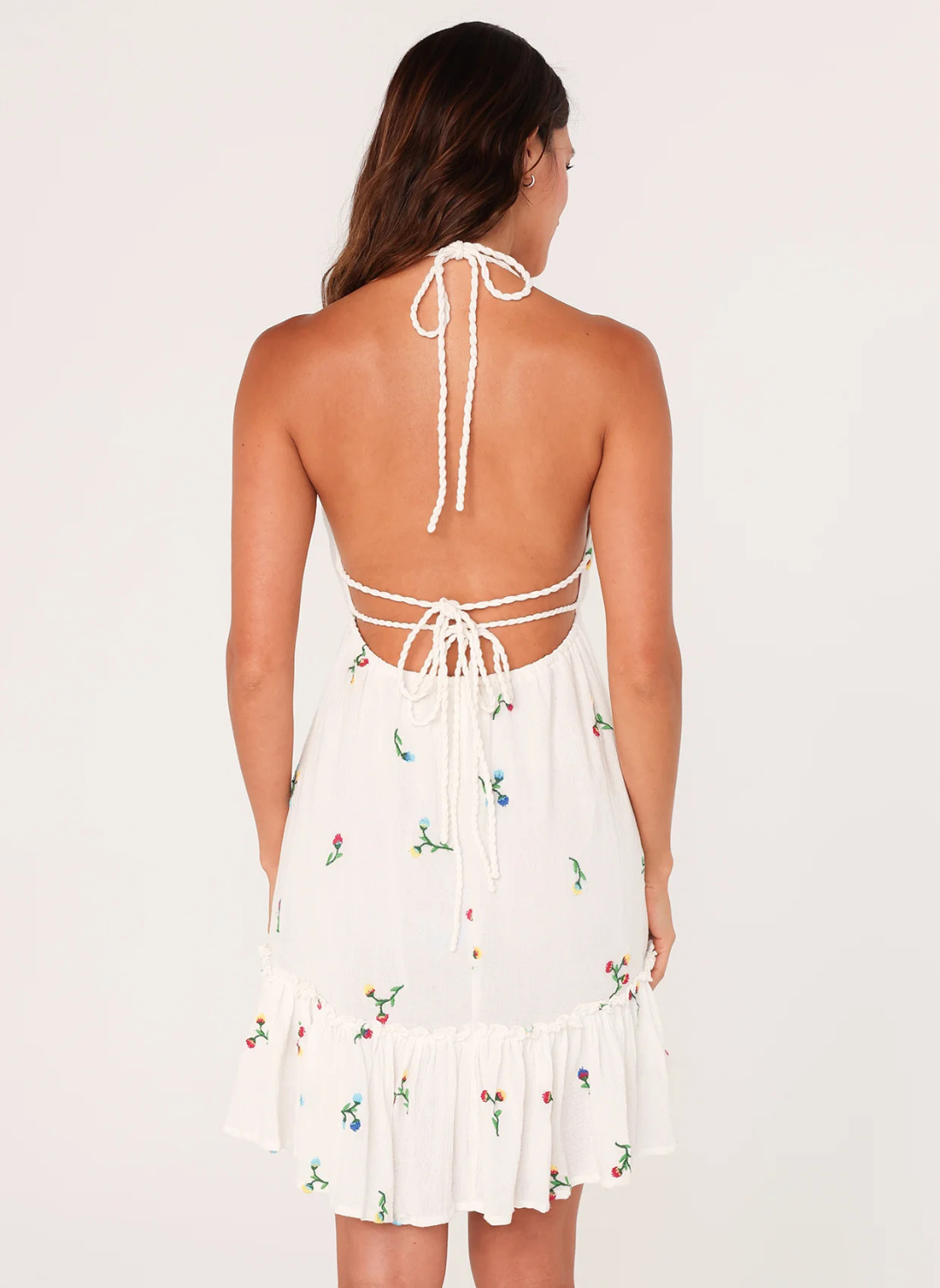 Back view of a woman wearing a white mini dress with floral embroidery, deep V-neckline, braided halter straps, and open tie back—featuring a ruffle hem and lined gauze fabric for spring and summer wear.
