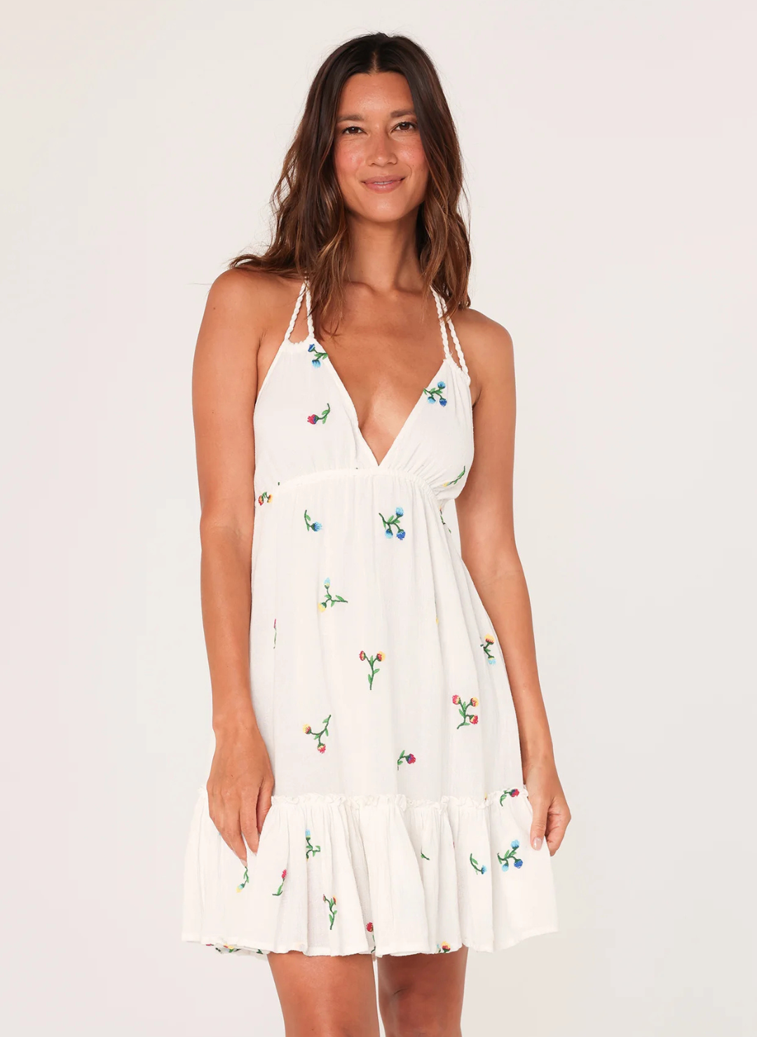 Woman wearing a white mini dress with floral embroidery, deep V-neckline, braided halter straps, and open tie back—featuring a ruffle hem and lined gauze fabric for spring and summer wear.