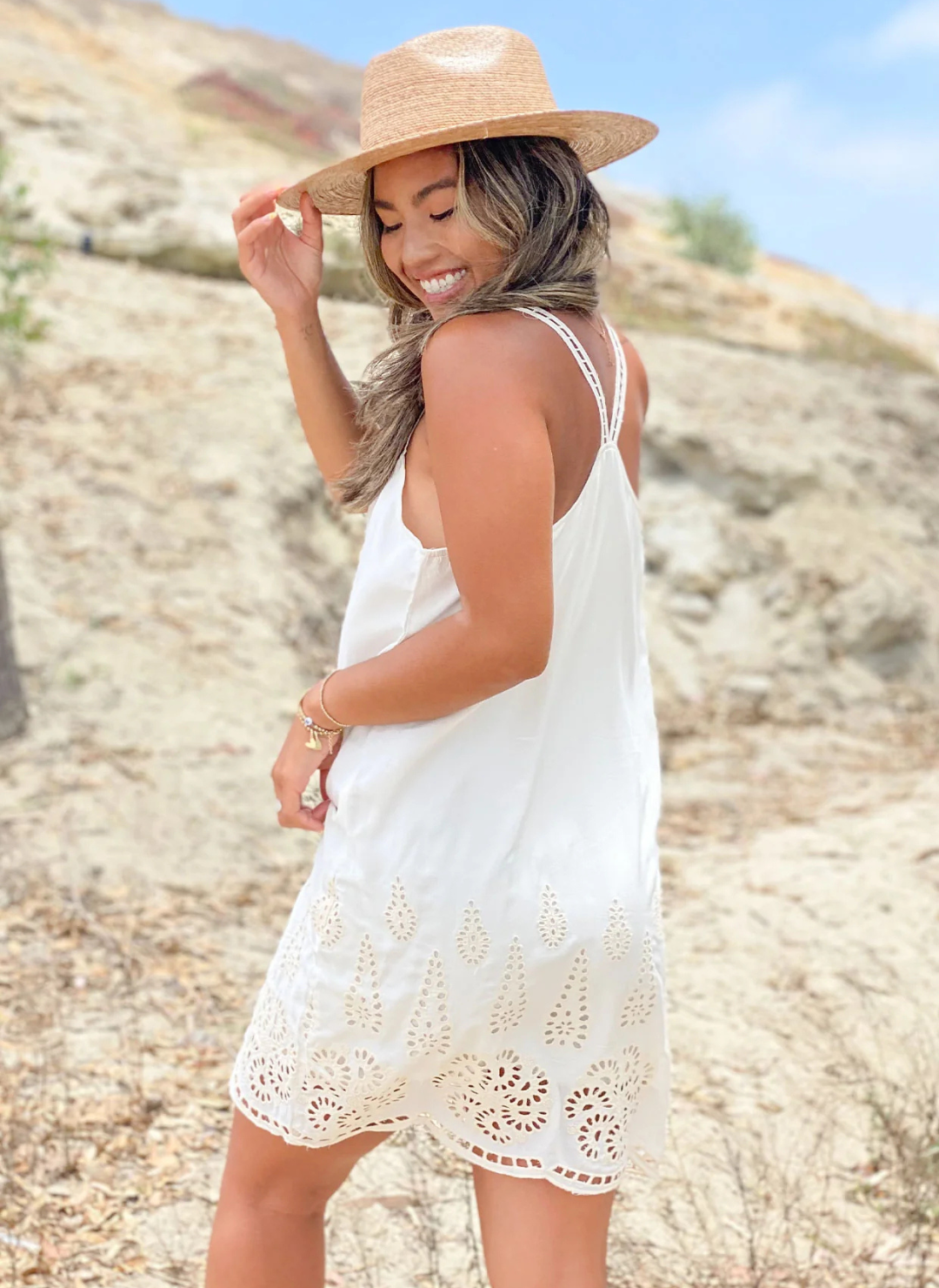 Woman wearing a white mini dress with eyelet embroidery along the hem, scoop neckline, and double strap detail—lightweight and perfect for beach days or casual summer outings.