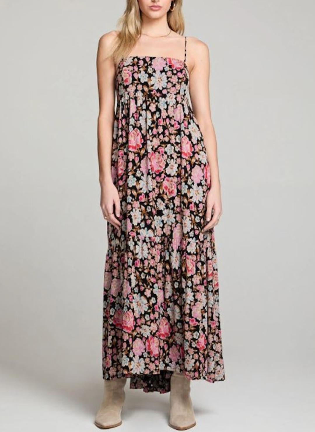 Woman wearing a black maxi dress with pink and white floral print, spaghetti straps, tiered skirt, and a bow tie-back detail. Styled with boots and a relaxed, summery feel.
