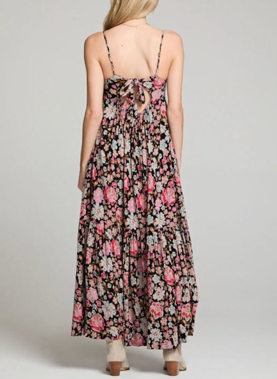 Back view of a woman wearing a black maxi dress with pink and white floral print, spaghetti straps, tiered skirt, and a bow tie-back detail. Styled with boots and a relaxed, summery feel.