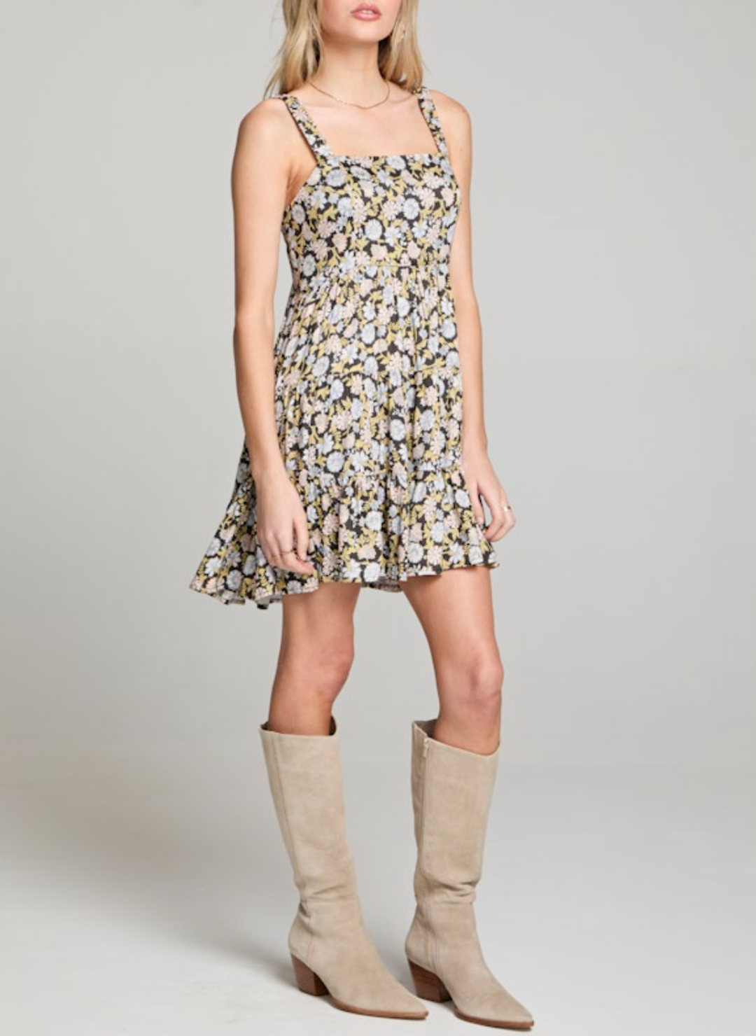 Woman wearing a black floral mini dress with square neckline, wide straps, tiered skirt, and back cutout with tie. Styled with tall beige boots for a modern western look.
