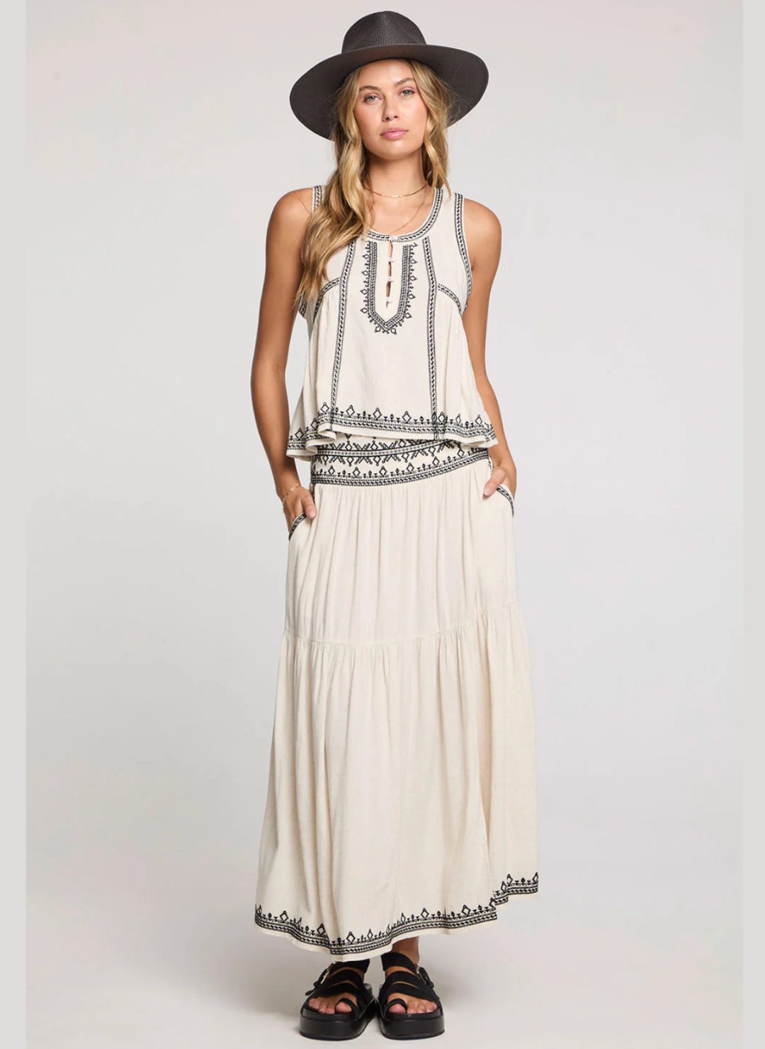 Woman wearing a long white dress with black patterns and a hat on a plain background