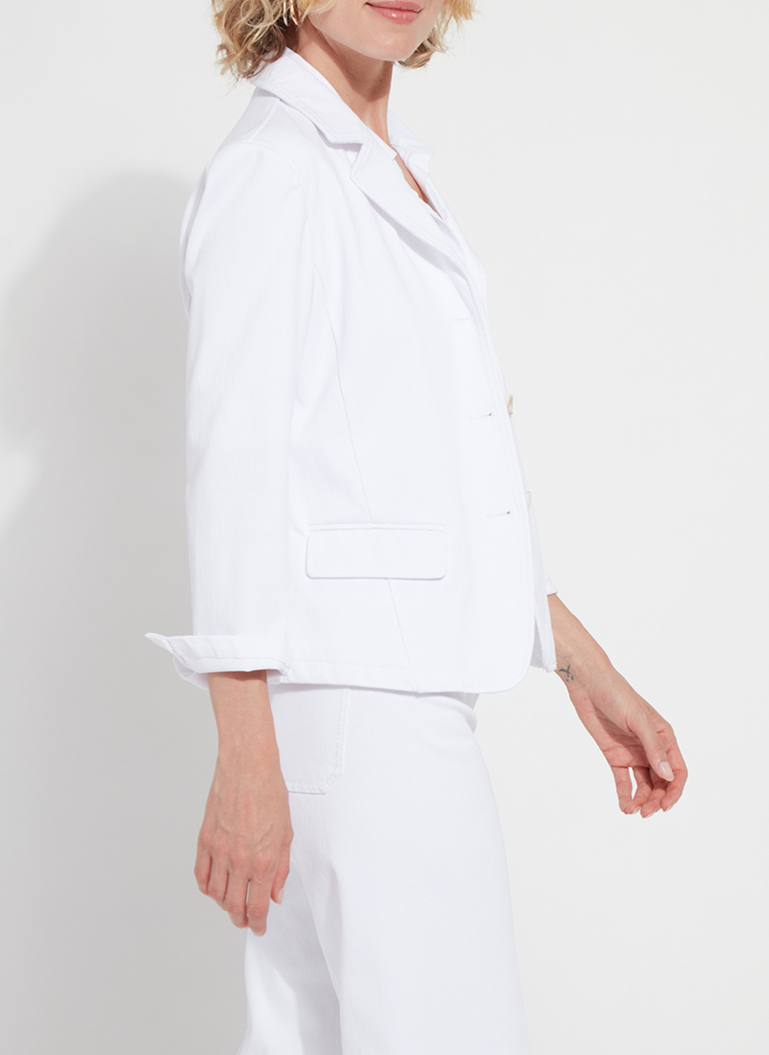 Side view of a woman wearing a crisp white Lyssé Della Stretch Denim Blazer with rolled sleeves, notched lapels, and front button closure. This tailored yet stretchy blazer is styled with matching white denim, offering a clean, polished boutique look perfect for work or weekend wear.