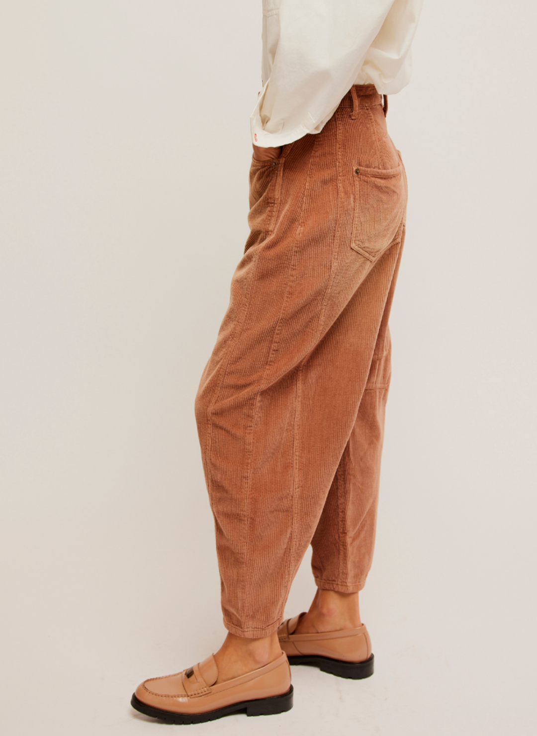 Side View: Pictured a corduroy pant in a rust color. Darts at the inner knee and a tapered hem. White shirt paired with it and loafers.