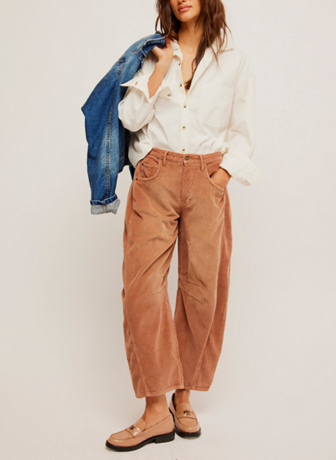 Front view of model holding a jean jacket on her shoulder. Pictured a corduroy pant in a rust color. Darts at the inner knee and a tapered hem. White shirt paired with it and loafers.