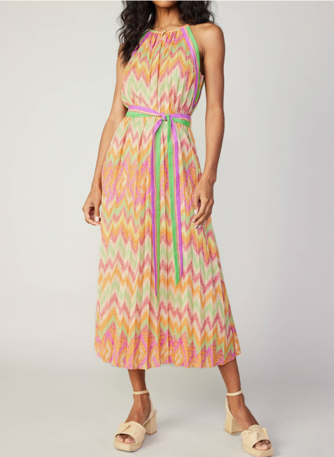 Model wearing colorful chevron pleated dress with high neckline, belt tie, and neckline button closure.