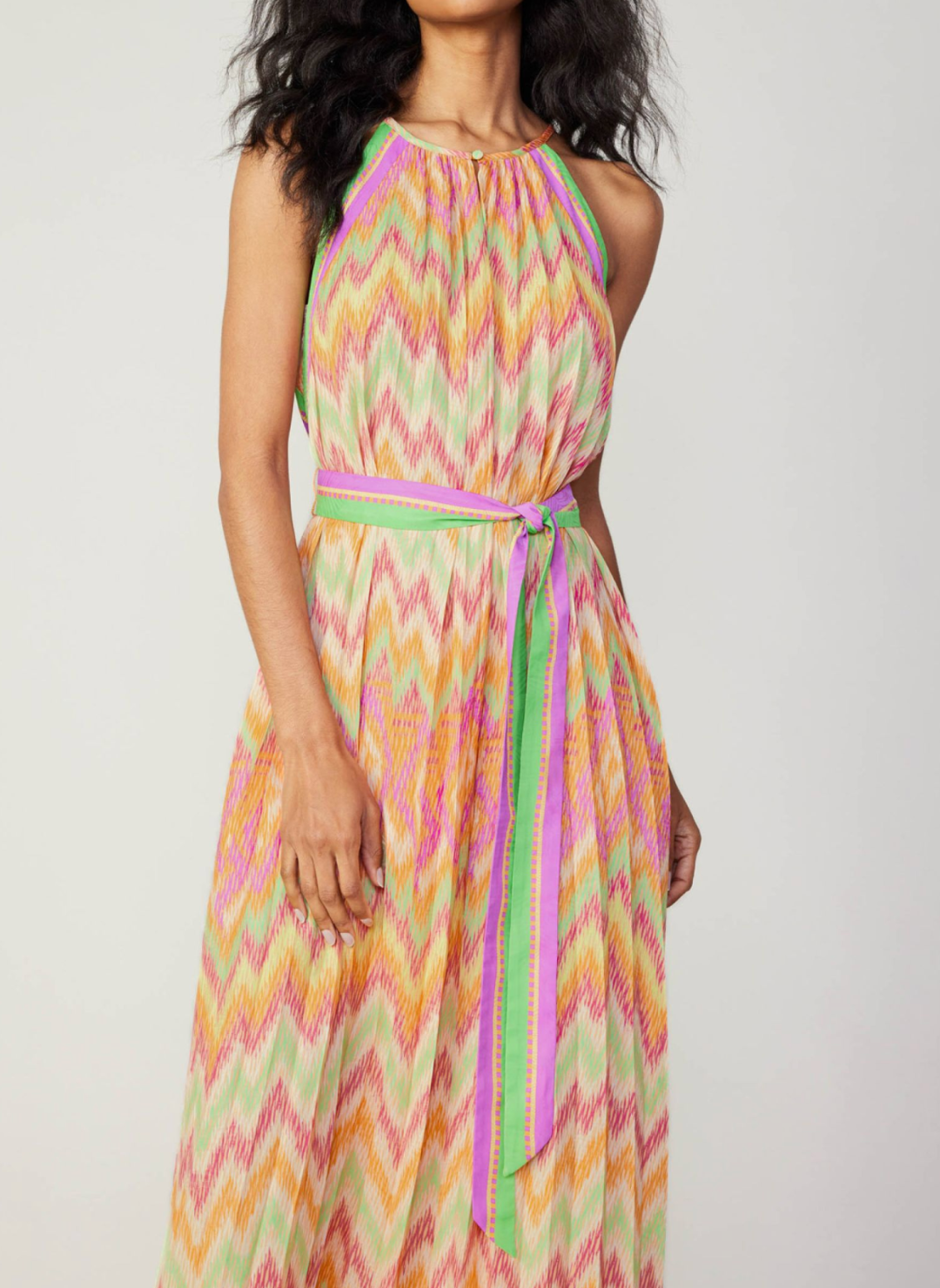 Model wearing colorful chevron pleated dress with high neckline, belt tie, and neckline button closure.