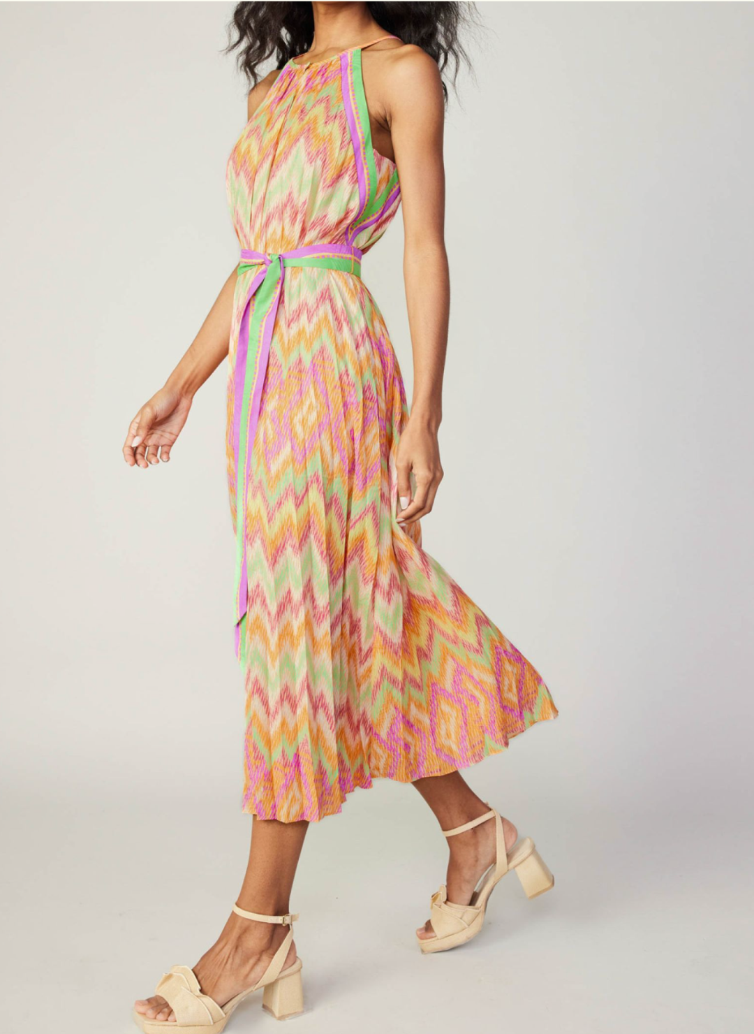 Side walking view of model wearing colorful chevron pleated dress with high neckline, belt tie, and neckline button closure.