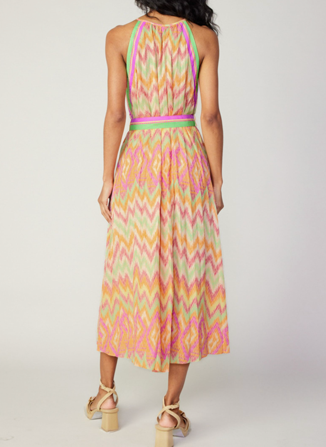 Back view of model wearing colorful chevron pleated dress with high neckline, belt tie, and neckline button closure.