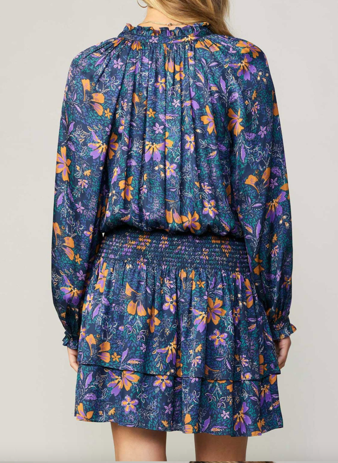 Back view of model wearing blue long sleeve dress included with smocked waist, ruffled neckline, and floral print.