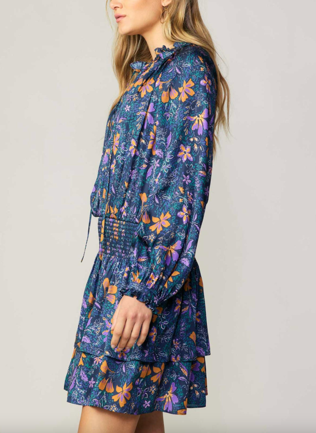 Side view of model wearing blue long sleeve dress included with smocked waist, ruffled neckline, and floral print.