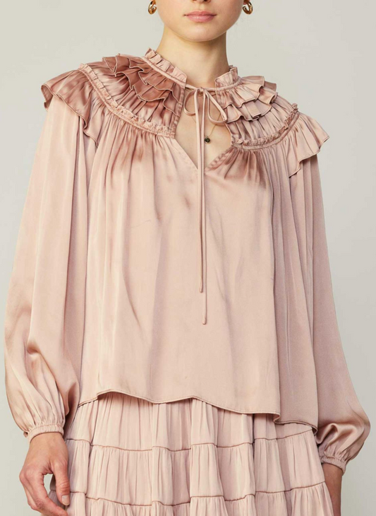 Model wearing light pink satin blouse with ruffles and front neckline tie. 