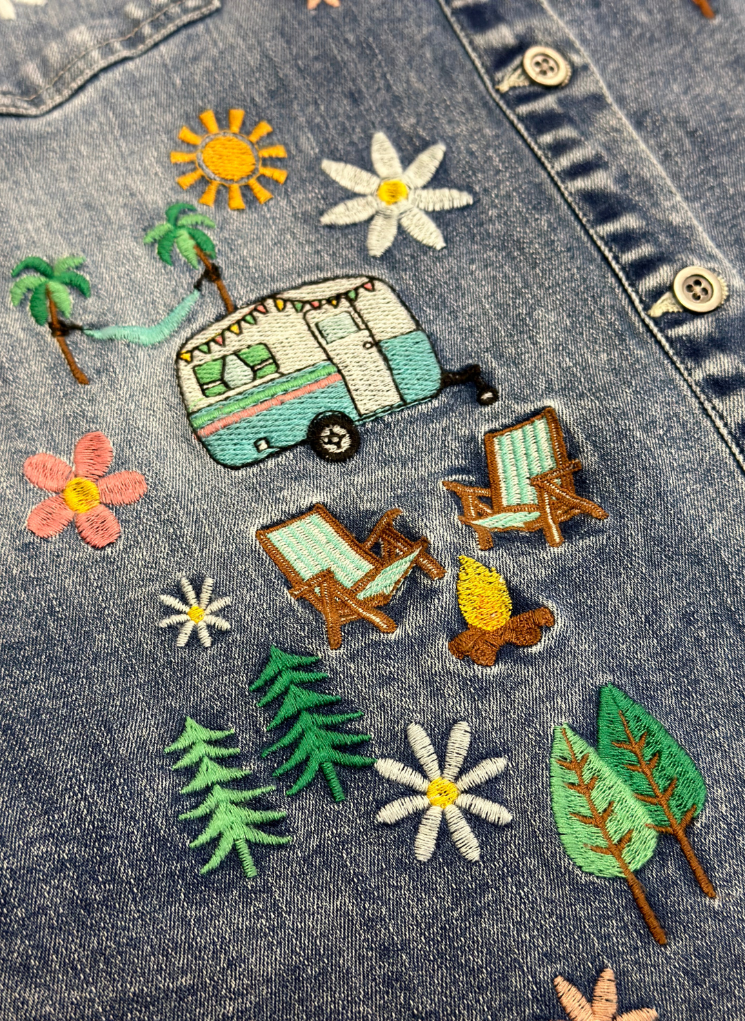 Close up flat view of denim camper shirt with embroidered, trailer, sun, hammock, campfire, chairs, trees, and flowers.