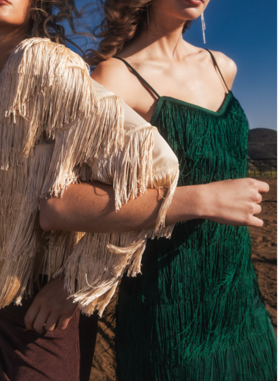 Model wearing green Gi Fringe Strap Dress is linked arms with another model wearing a cream colored fringe outfit. 