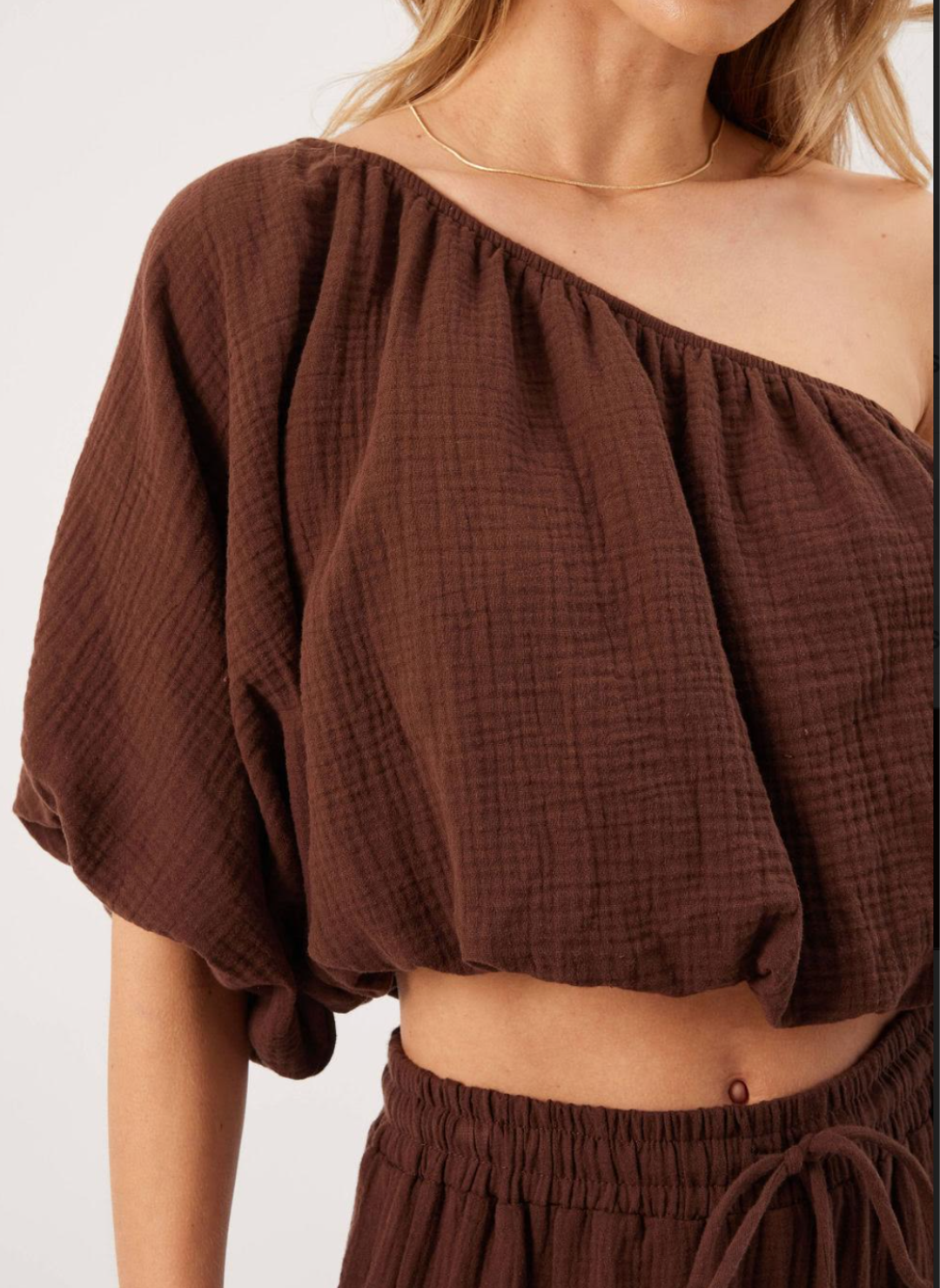 Close up view of model wearing brown KP Lina Top with one shoulder detail and bubble hem along with matching pants.