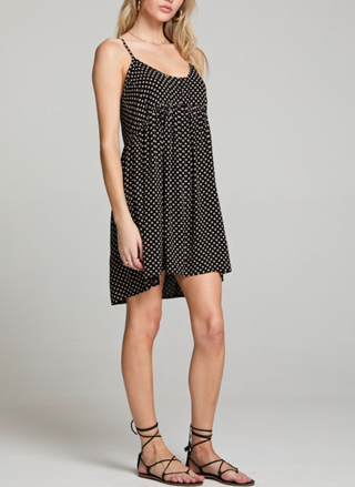 Woman wearing a black spaghetti strap mini dress with beige polka dots, racerback detail, and a flowy babydoll silhouette. Styled with lace-up sandals for a casual warm-weather look.