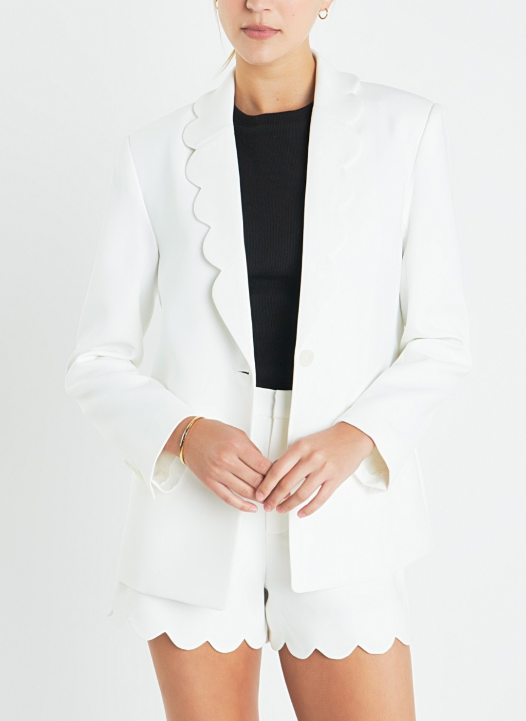 Front view of model wearing white Sassy Scalloped Blazer with black tank top underneath and matching white scalloped shorts. Hands are together in front. White background.