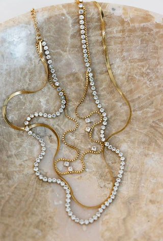 A gold-toned double-strand necklace with clear rhinestone and cubic zirconia embellishments, displayed on a textured beige stone surface.