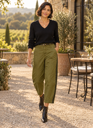 A woman walking outdoors on a gravel patio wearing olive green cropped pants with black side stripes, a black long-sleeve top, and black ankle boots.