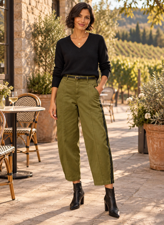 A woman stands outdoors on a stone patio in front of a vineyard, wearing olive green wide-leg cropped pants with dark side stripes, a black long-sleeve V-neck top, and black ankle boots.