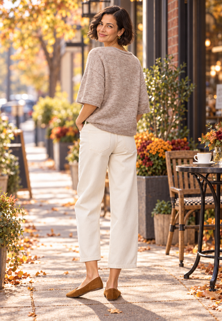 A woman stands on an outdoor sidewalk in an autumn street setting, wearing a loose beige knit top, off-white cropped pants, and tan flats.