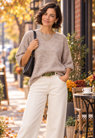 A woman stands outdoors on a city sidewalk wearing a loose heathered taupe knit sweater, white wide-leg pants, a green belt, and carrying a black shoulder bag.