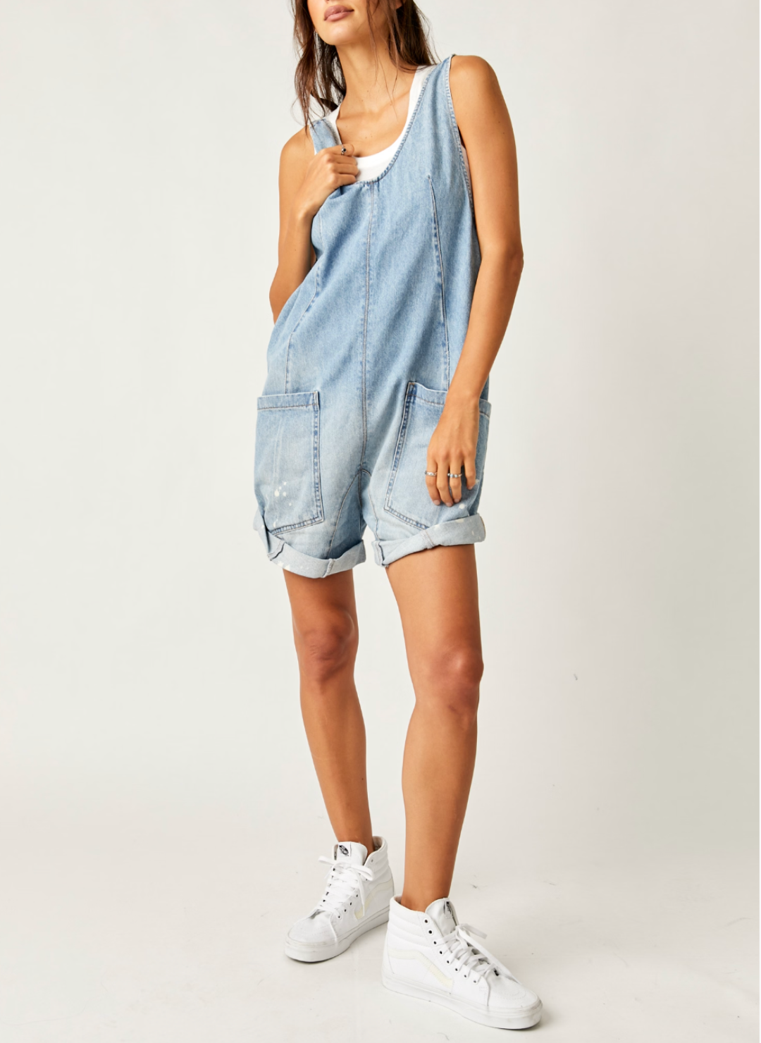 Model is wearing shoes denim romper with one hand on the strap portraying the relaxed fit. 