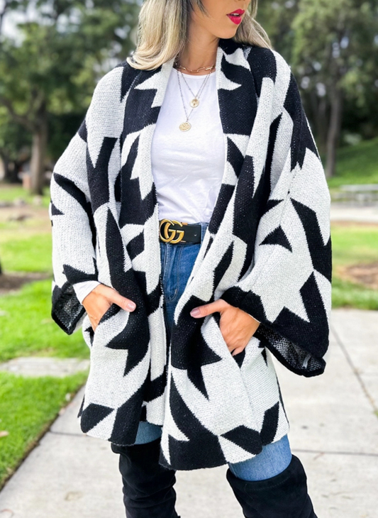 Full front view of model wearing the Houndstooth Oversized Cardi paired with a white top, denim bottoms, and black boots. Model is standing outside. 