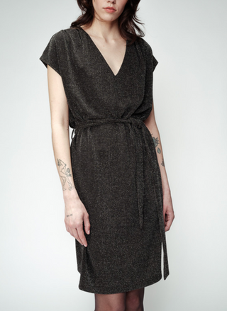 Front view of Model wearing the Bright Lights Belted Dress that ties in the towards the right side on the waist. The short sleeve dress has V-neck line. Dress is paired either black tights. Models arms are by her side. There is a white background.