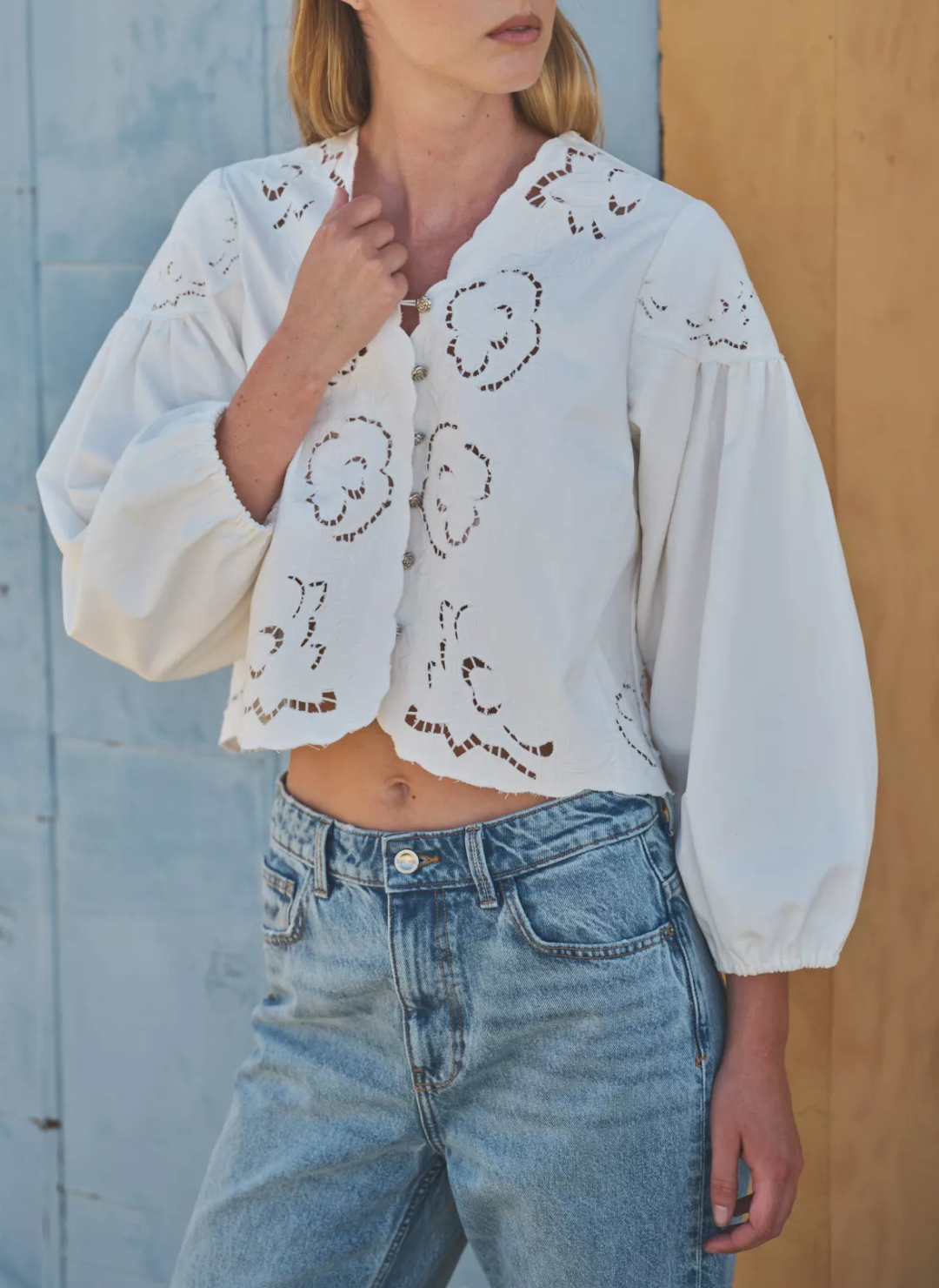 Model is wearing embroidered cream colored blouse with scalloped neckline. Models left hand is holding on to the blouse portraying the neckline.