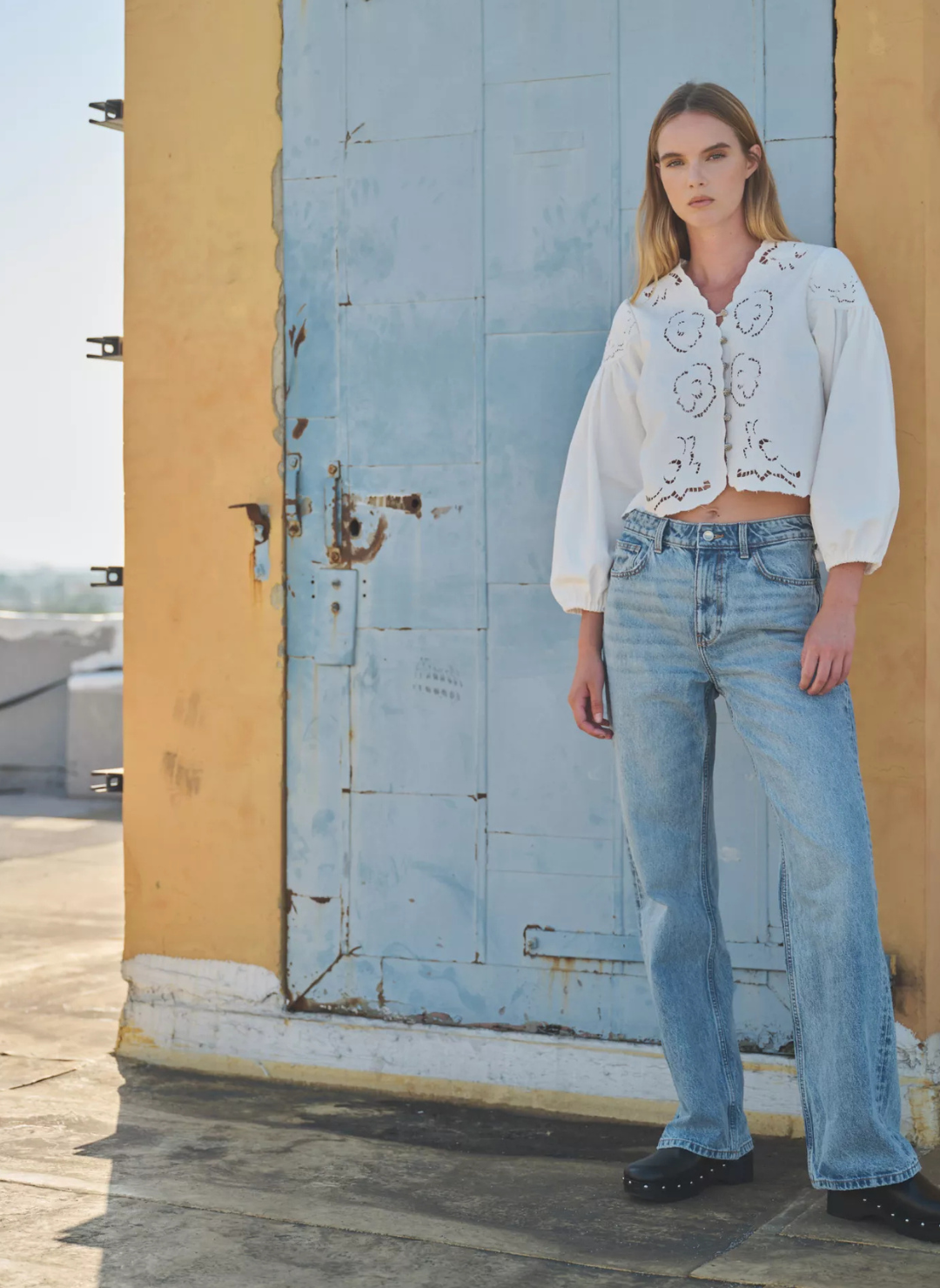 Model is standing farther away wearing embroidered cream colored blouse with denim jeans. Model is standing in front of blue door and yellow wall.