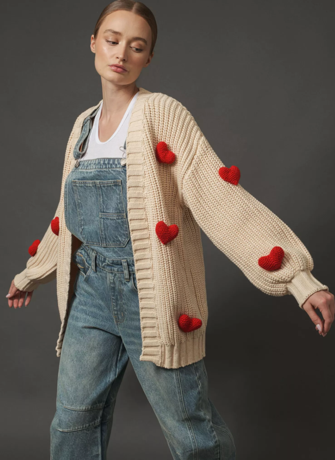 Model is wearing an oversized cream cardigan with red puffy hearts. Model is wearing overalls and a white t-shirt underneath the cardigan.