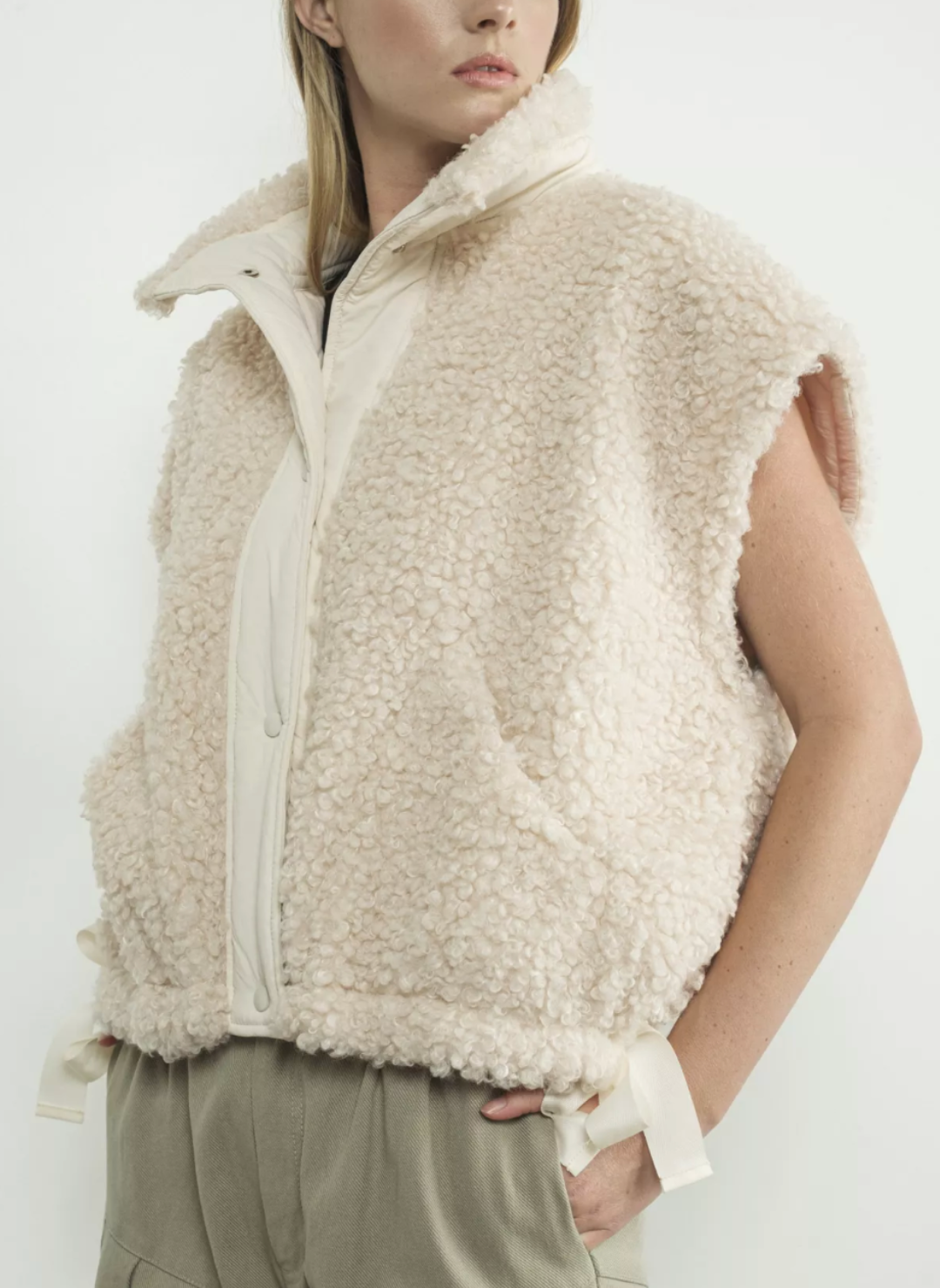 Front/side view of fuzzy snow colored button down vest. There are tie bows on each side at the bottom of the vest. Models hands are in pockets of light green pants. White background.