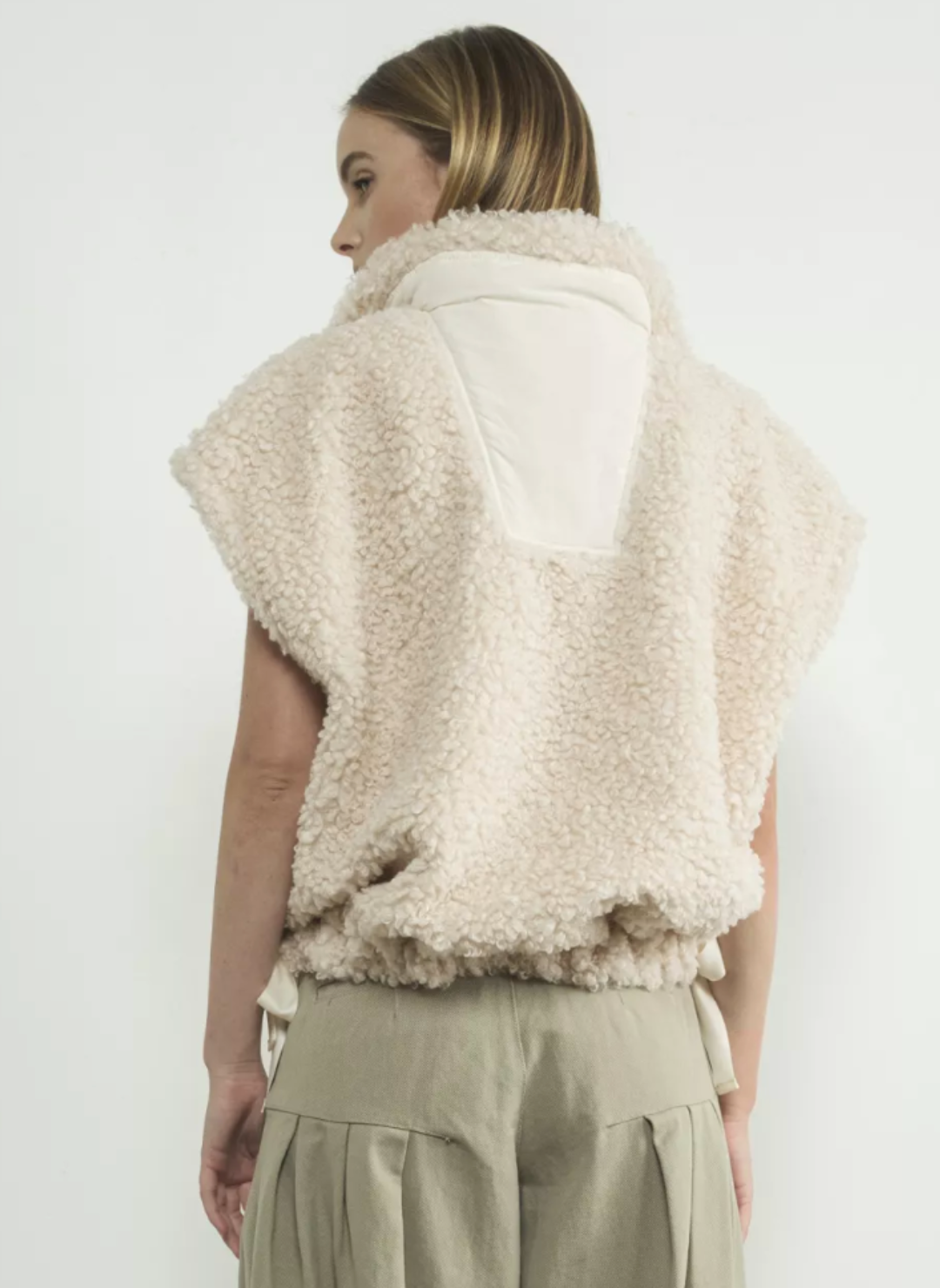 Back view of snow colored fuzzy vest paired with light green pants. There is a middle part of the vest with a smooth material. Models hands are by her side. White background.