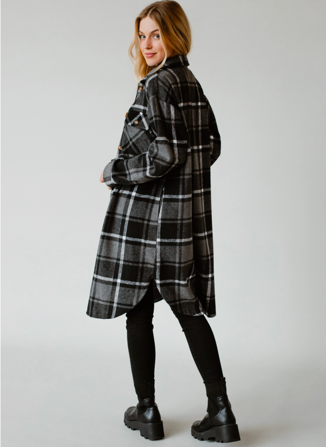 Model is slight turned portraying the backside in the plaid coat. Model is also wearing black pants with back boots. White background.