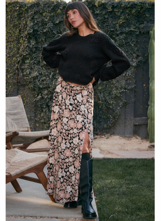 Model wearing floral maxi skirt with a slit at the leg.  Wearing black boots & black sweater in an outdoor setting.