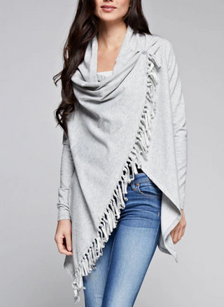 Woman wearing a light grey fringe wrap sweater with snap closure at the shoulder, styled over jeans and a tank top. Cozy and convertible boutique layering piece.