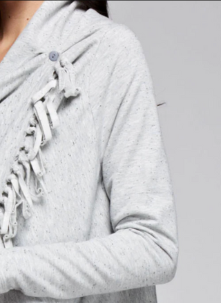 Woman wearing a light grey fringe wrap sweater with snap closure at the shoulder, styled over jeans and a tank top. Cozy and convertible boutique layering piece.
