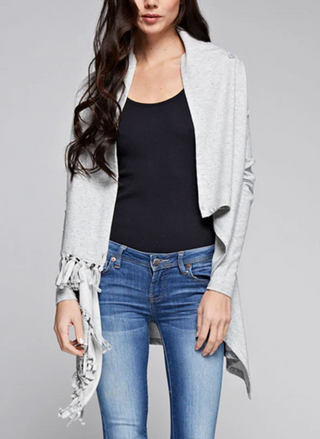Woman wearing a light grey fringe wrap sweater with snap closure at the shoulder, styled over jeans and a tank top. Cozy and convertible boutique layering piece.