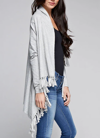 Woman wearing a light grey fringe wrap sweater with snap closure at the shoulder, styled over jeans and a tank top. Cozy and convertible boutique layering piece.