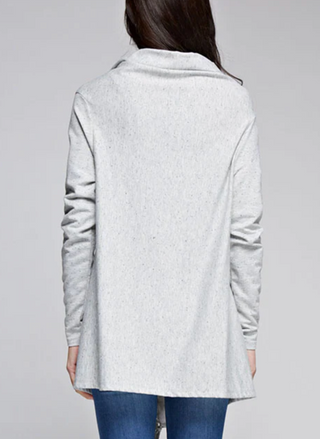 Back view of woman wearing a light grey fringe wrap sweater with snap closure at the shoulder, styled over jeans and a tank top. Cozy and convertible boutique layering piece.