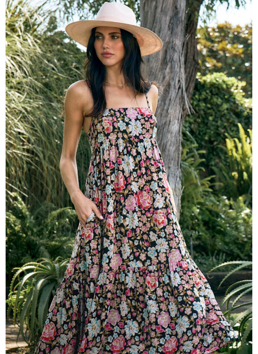 Woman wearing a black maxi dress with pink and white floral print, spaghetti straps, tiered skirt, and a bow tie-back detail. Styled with a sun hat and a relaxed, summery feel.