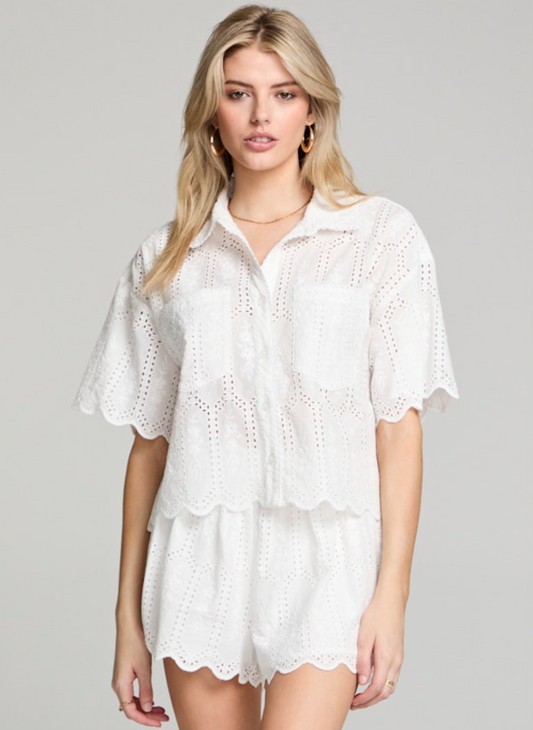 Woman wearing a cropped white cotton eyelet shirt with scalloped hems, front chest pockets, and flutter short sleeves. Styled buttoned up with matching white eyelet shorts.