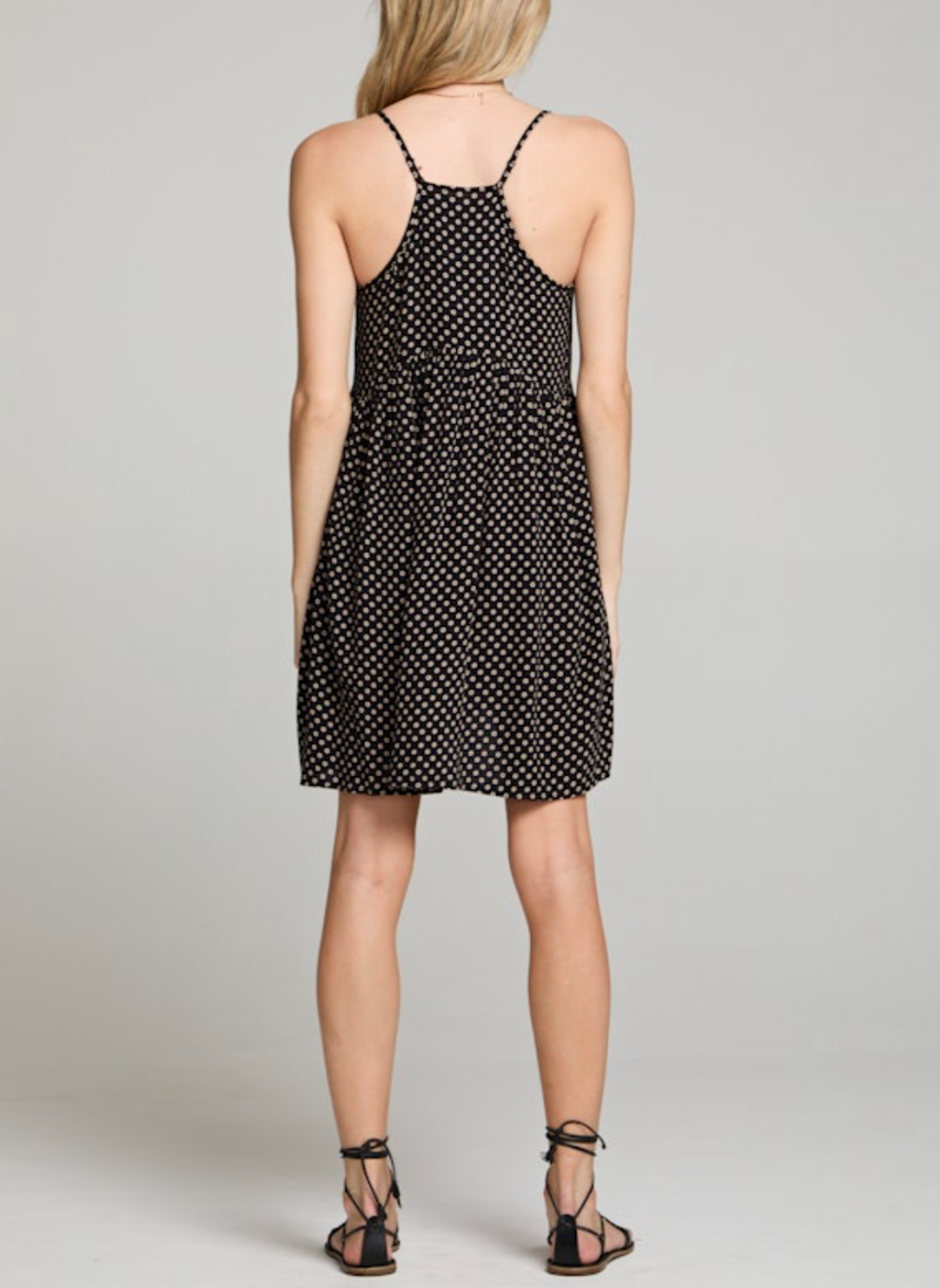 Woman wearing a black spaghetti strap mini dress with beige polka dots, racerback detail, and a flowy babydoll silhouette. Styled with lace-up sandals for a casual warm-weather look.