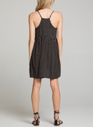 Woman wearing a black spaghetti strap mini dress with beige polka dots, racerback detail, and a flowy babydoll silhouette. Styled with lace-up sandals for a casual warm-weather look.